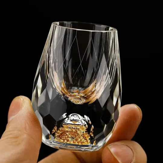 a hand holding a luxurious gold gemstone shot glass. The glass features a hand-blown faceted design with a shimmering gold-infused base, showcasing its 0.5 oz capacity. This elegant piece is part of an exclusive luxury drinkware collection, highlighting high-quality craftsmanship and unique style.