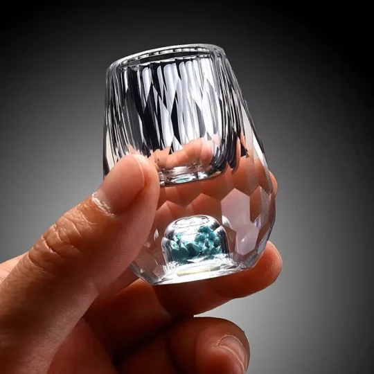 a hand holding a luxurious turquoise gemstone shot glass. The glass features a hand-blown faceted design with a vibrant turquoise-infused base, showcasing its 0.5 oz capacity. This elegant piece is part of an exclusive luxury drinkware collection, highlighting high-quality craftsmanship and unique style.