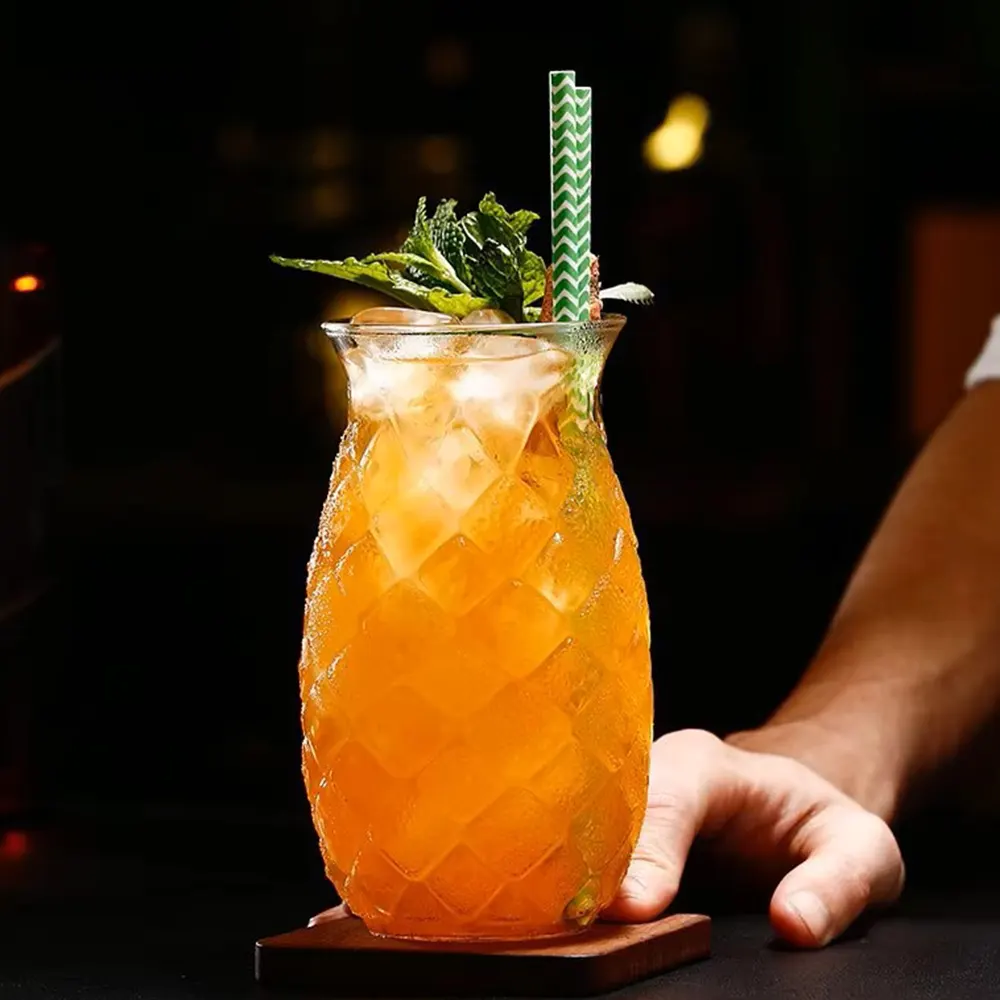 Close-up of a hand holding a textured glass with an orange drink, ice cubes, mint leaves, a slice of lime or lemon, and a striped straw, against a dark background.