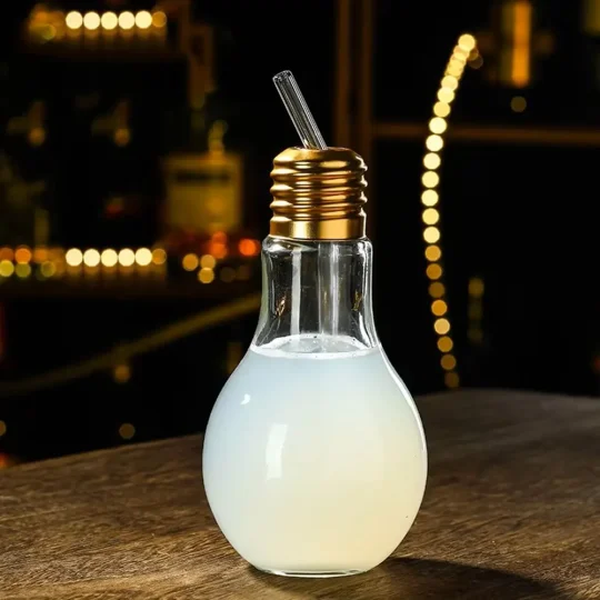 Unique light bulb-shaped cocktail glass filled with a milky beverage, featuring a metal screw cap and a straw, set on a dark bar with soft bokeh lights in the background.