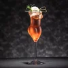 200ml La Belle Femme stemmed glass shaped like a woman’s body, filled with a rose-colored drink, garnished with rosemary and an ice sphere, set against a dark background.