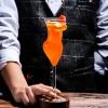 Person holding a 200ml La Belle Femme stemmed glass with a woman’s silhouette, filled with an orange cocktail, garnished with a citrus slice and cherry, set on a wooden bar counter.