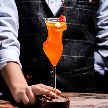 Person holding a 200ml La Belle Femme stemmed glass with a woman’s silhouette, filled with an orange cocktail, garnished with a citrus slice and cherry, set on a wooden bar counter.