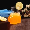 Novelty ‘La Derrière’ glass filled with orange beverage, ice cubes, and a lemon slice on the rim, placed on a brown surface with a blurred background featuring a blue book and purple flowers – ideal for fun drinkware and cocktail lovers.