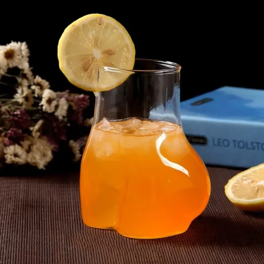 Novelty ‘La Derrière’ glass filled with orange beverage, ice cubes, and a lemon slice on the rim, placed on a brown surface with a blurred background featuring a blue book and purple flowers – ideal for fun drinkware and cocktail lovers.