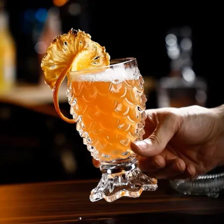 Close-up of a hand holding an ornate Mermaid’s Embrace cocktail glass filled with amber liquid and garnished with a dehydrated citrus wheel.