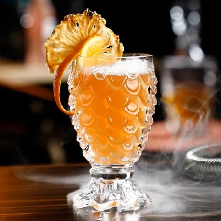 Elegant Mermaid’s Embrace cocktail glass with shimmering scale-like design filled with amber drink garnished by dehydrated orange.