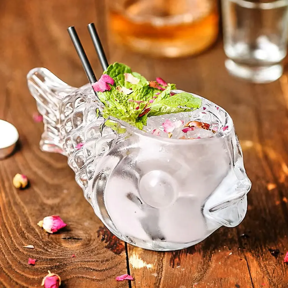 A fish-shaped cocktail glass named Mr. Fishy, filled with a clear liquid, garnished with green leaves and pink petals, with two black straws, set on a wooden surface with a blurred bottle and glass in the background.