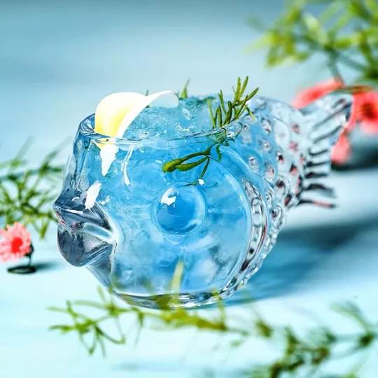 A whimsical fish-shaped cocktail glass named Mr. Fishy, filled with a blue cocktail and garnished with a lemon twist and rosemary sprig, set against a soft blue background with floral accents.