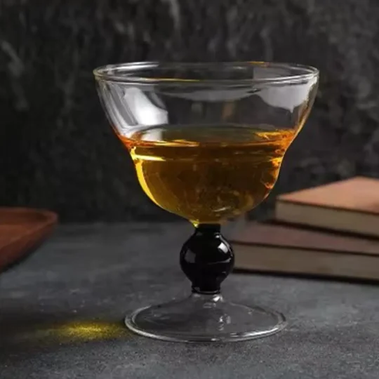 A close-up of the Obsidian Sphere 5.75 oz Cocktail Glass featuring a wide bowl and a distinctive black-beaded stem, placed on a dark surface with books in the background.