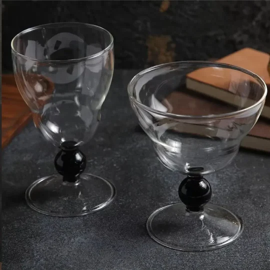 One Obsidian Sphere Whisky Glass with elongated shape and black-beaded stem next to one Obsidian Sphere Cocktail Glass with wide bowl on a dark stone countertop.