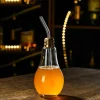 Light bulb-shaped glass filled with an orange drink and a metal straw, placed on a dark surface with a moody background.