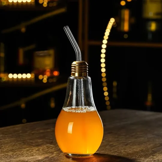 Light bulb-shaped glass filled with an orange drink and a metal straw, placed on a dark surface with a moody background.