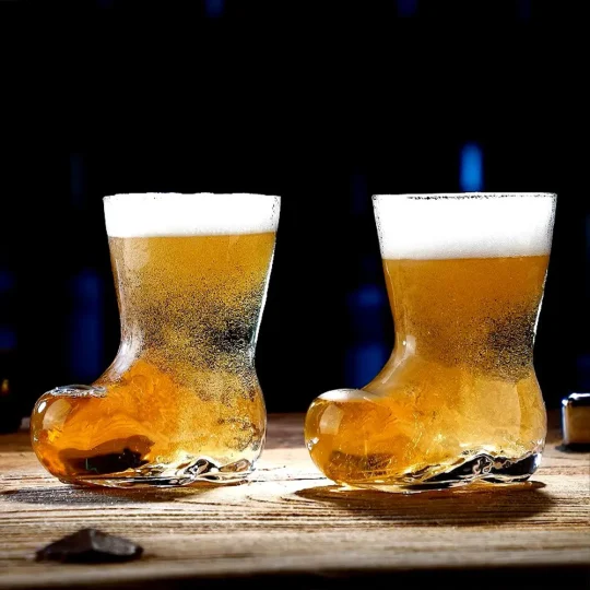 Two cowboy boot-shaped glasses filled with golden beer, placed on a wooden surface with a dark background, highlighting the bubbles and amber color of the beer.