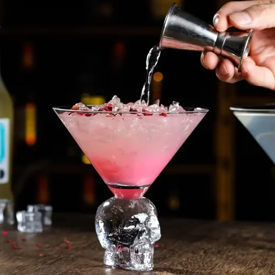 Pink cocktail being poured into a large martini glass with a skull-shaped stem, filled with ice and garnished with red particles.