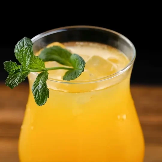 Close-up of a curved cocktail glass filled with a yellow-orange beverage, ice cubes, and garnished with a sprig of mint, set against a dark background.