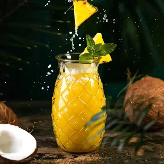 Textured glass resembling pineapple skin, filled with a yellow tropical drink, garnished with mint, and splashing pineapple pieces, with coconuts in the background.