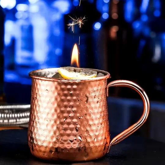 Close-up of a hammered texture copper-plated stainless steel Moscow Mule mug with ice, lemon garnish, and a lit sparkler, 500 ml capacity.