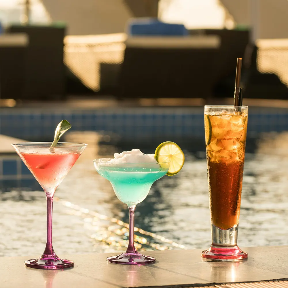 Three different cocktails placed near a swimming pool at sunset, showcasing the elegance and style of premium drinkware designed to enhance classic cocktails like martinis, margaritas, and daiquiris.