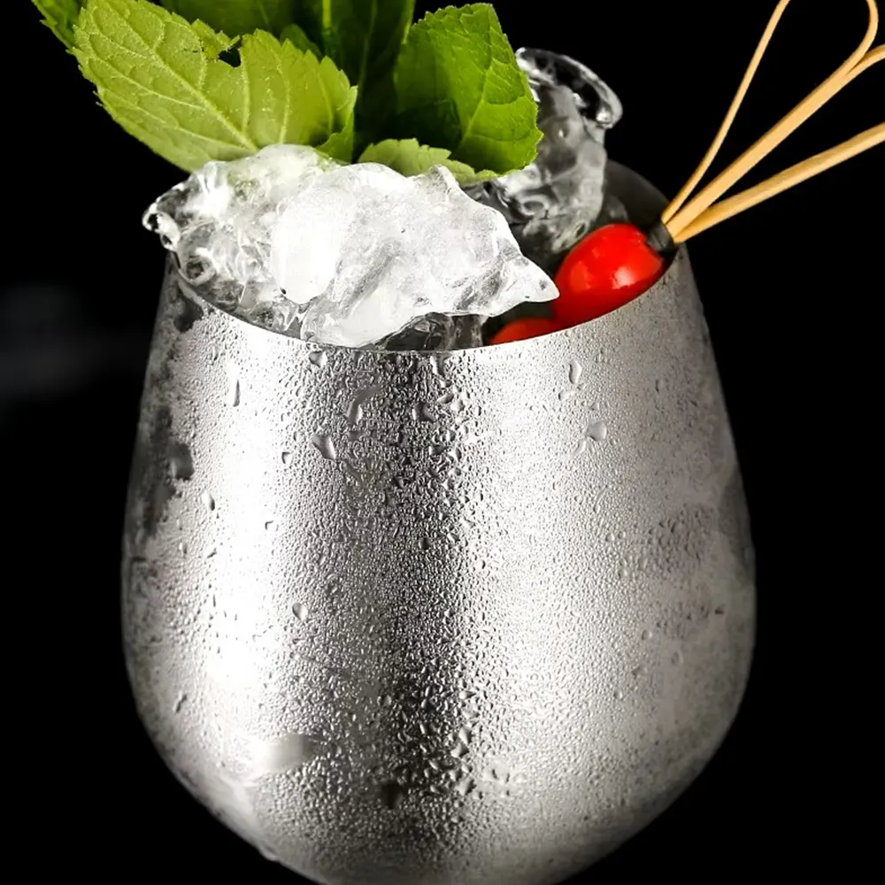 Close-up of a silver metallic wine glass with a smooth finish, filled with ice and garnished with mint leaves and a cherry, set against a dark background.