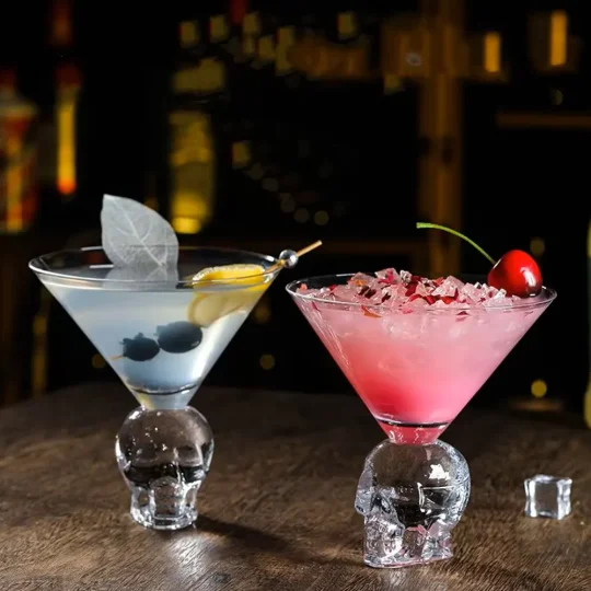 Two cocktails in Skulltini Glasses with skull-shaped stems, one clear with a lemon twist and blueberries, the other pink with crushed ice and a cherry, set in a dimly lit bar.