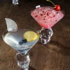 Two cocktails in Skulltini Glasses with skull-shaped stems, one garnished with a lemon slice and blueberries, the other with a cherry, placed on a wooden surface.