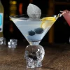 Clear martini served in an innovative Skulltini Glass with skull-shaped stem, garnished with olives and lemon twist on a bar counter.