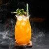 Textured glass with an orange tropical drink, garnished with green leaves and a cinnamon stick, surrounded by misty smoke, placed on a wooden coaster.