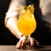 Hand holding a stemmed curved cocktail glass filled with a yellow-orange drink, garnished with a lime slice and mint leaves, against a dark background.