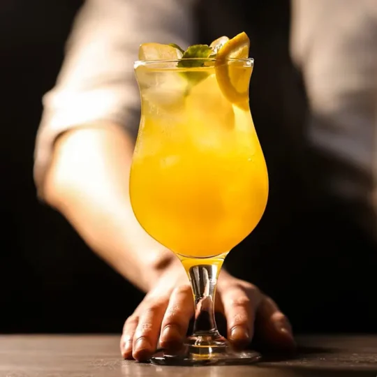 Hand holding a stemmed curved cocktail glass filled with a yellow-orange drink, garnished with a lime slice and mint leaves, against a dark background.