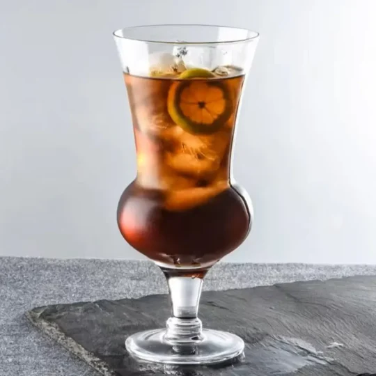 A 660 ml round belly cocktail glass filled with a dark-colored iced beverage, garnished with a slice of lemon, placed on a grey stone surface.