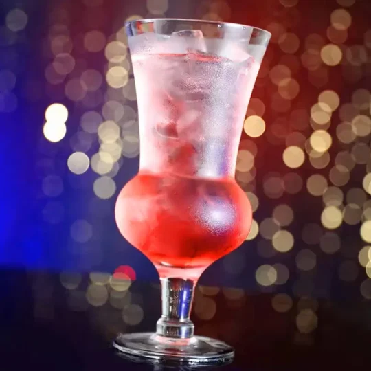 A 660 ml round belly cocktail glass filled with a red and clear liquid cocktail, with ice cubes, placed against a background with colorful bokeh lights.