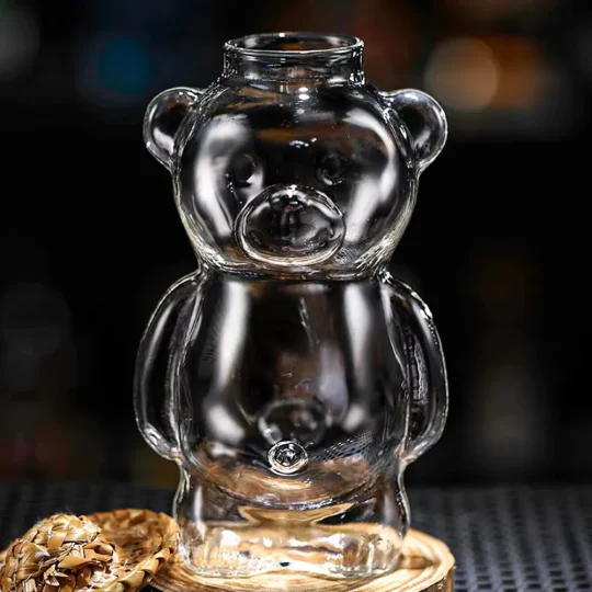 Empty bear-shaped cocktail glass, ‘The Honey Bear,’ with a screw top lid, placed on a bar or kitchen counter next to a pine cone, highlighting its unique design and clear glass material.