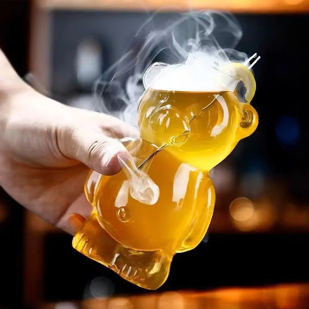 Hand holding ‘The Honey Bear’ cocktail glass filled with a golden liquid, with steam rising from the top, showcasing the bear-shaped design and spout for pouring.