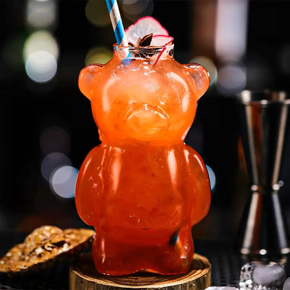 Bear-shaped cocktail glass, ‘The Honey Bear,’ filled with an orange drink, garnished with a star anise pod and cherry, with a blue and white striped straw, placed on a wooden coaster.