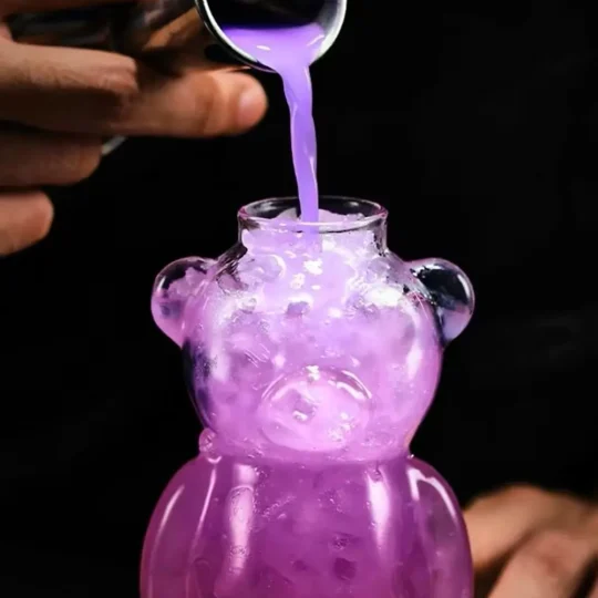 Close-up of ‘The Honey Bear’ cocktail glass being filled with a bubbling purple liquid from a metal ladle, highlighting the bear-shaped design and dynamic motion of the pour.