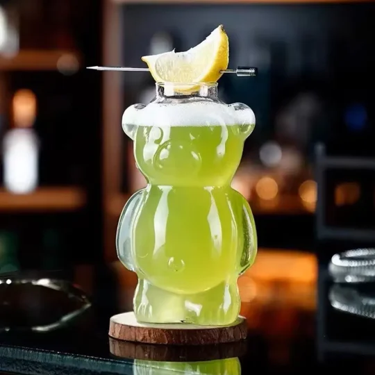 Bear-shaped cocktail glass, known as ‘The Honey Bear,’ with a 10.75 oz capacity, crafted from high borosilicate glass featuring a smooth design and thickened bottom for stability, filled with a yellow-green cocktail and garnished with a lemon slice on the rim.