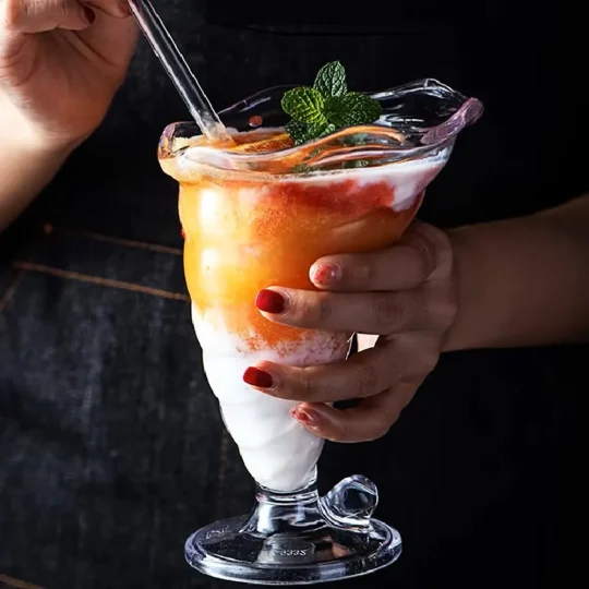 A person holding an eco-friendly cocktail glass with a spiral pattern, filled with a layered frozen dessert in orange and white, topped with mint.