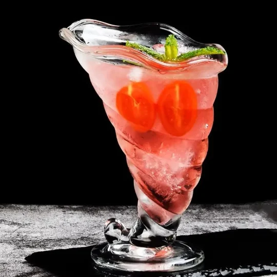 A uniquely designed, eco-friendly cocktail glass with a spiral pattern, filled with a pink cocktail and garnished with cherry tomatoes and green herbs.