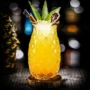 Elegant tall cocktail glass with diamond-etched design, filled with a vibrant yellow tropical drink, garnished with fresh green mint, a cinnamon stick, and a dehydrated citrus slice, presented on a wooden coaster.