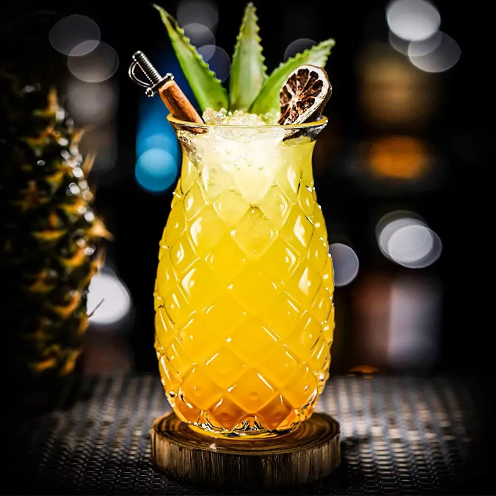 Elegant tall cocktail glass with diamond-etched design, filled with a vibrant yellow tropical drink, garnished with fresh green mint, a cinnamon stick, and a dehydrated citrus slice, presented on a wooden coaster.