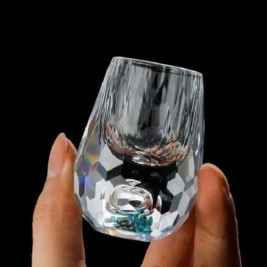 a hand holding a luxurious turquoise gemstone shot glass. The glass features a hand-blown faceted design with a vibrant turquoise-infused base, showcasing its 0.5 oz capacity. This elegant piece is part of an exclusive luxury drinkware collection, highlighting high-quality craftsmanship and unique style.