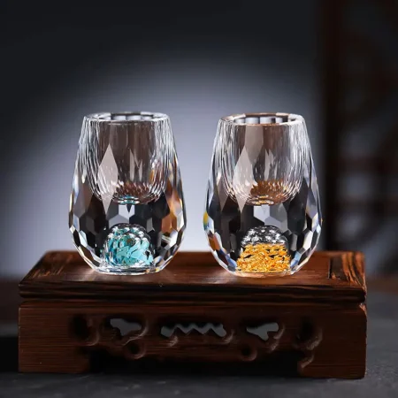 a pair of exquisite Gemstone shot glasses, one with a vibrant turquoise base and the other with a shimmering gold base, both featuring hand-blown glass with a unique faceted design that catches the light, set against a dark backdrop on an elegant wooden stand. Each glass boasts a 0.5 oz capacity, embodying sophistication and exclusivity in luxury drinkware.