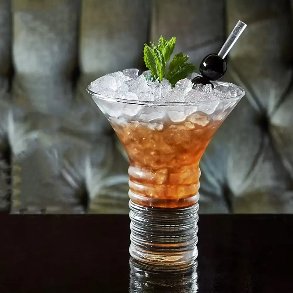 Twisted Martini Glass with an 8 oz capacity, featuring a spiral stem, filled with a dark cocktail garnished with a sprig of mint and a dark cherry, with a metal straw, set on a reflective surface.