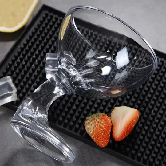 Clear cocktail glass with a spiral design, lying on its side with lemon slice and strawberries, surrounded by ice cubes on a textured black surface.