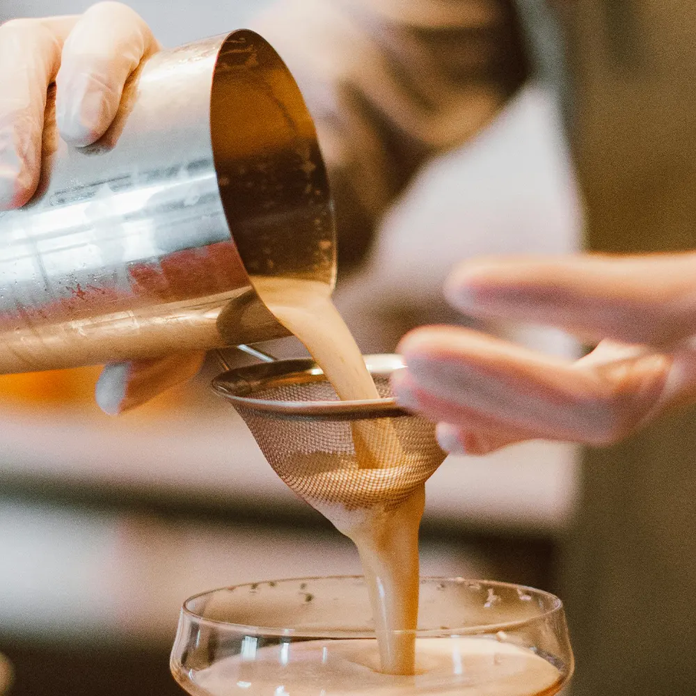 A close-up of a person pouring a liquid from a metal shaker through a strainer into a glass, highlighting the versatility and innovative design of premium drinkware for various beverages.