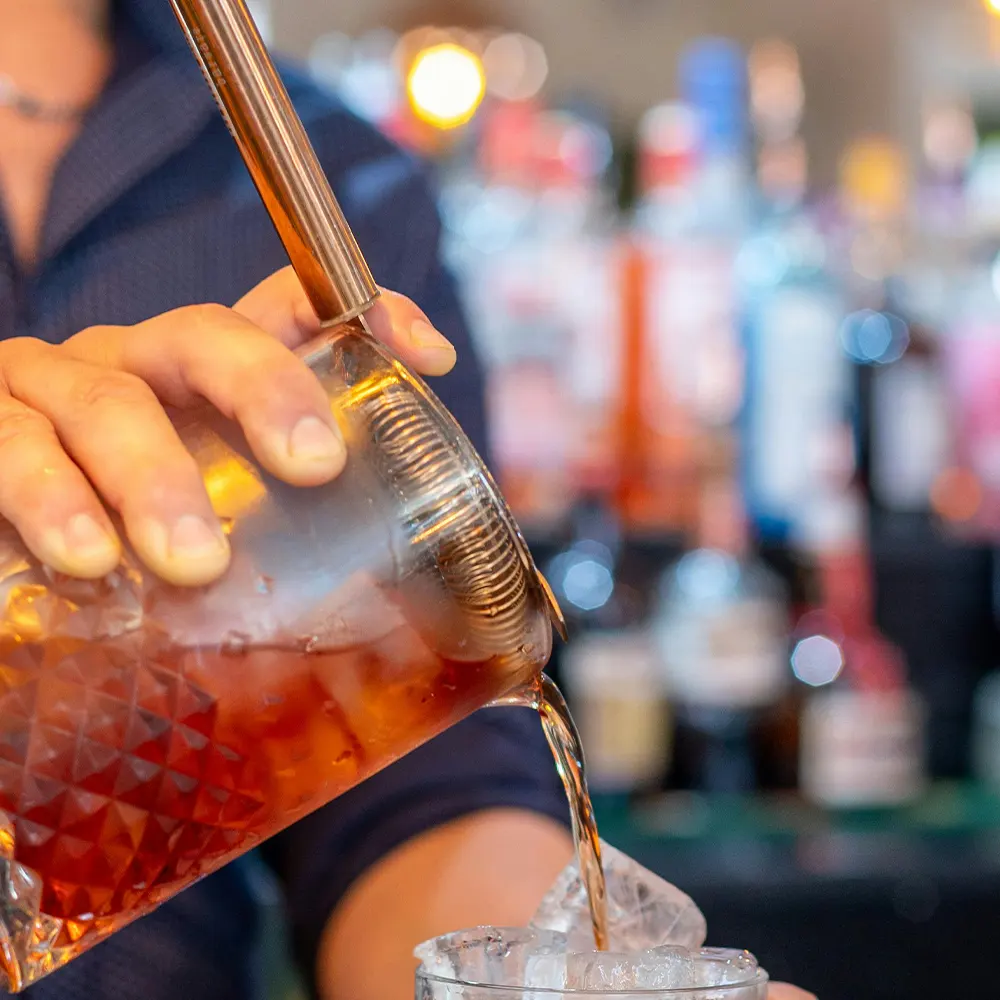 Close-up of hands pouring a brown liquid from a textured glass decanter into a glass filled with ice, highlighting the versatility of premium glassware for various beverages