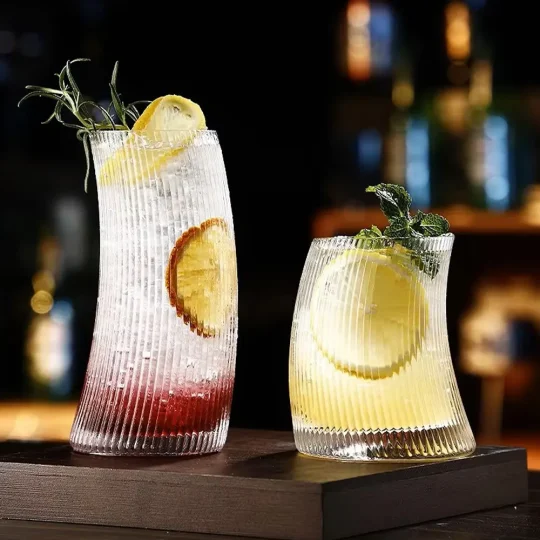 Two Vertical Elegance glasses with unique hunched design and vertical ridges, one filled with a red drink garnished with lemon and rosemary, and the other with a yellow drink garnished with lemon and mint.