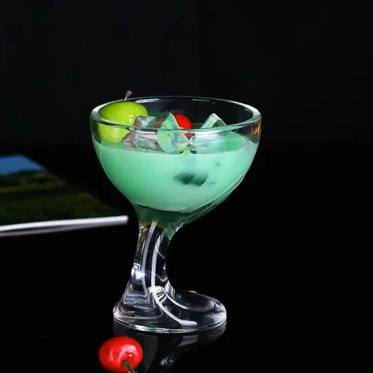 Clear cocktail glass with a spiral design, filled with green liquid, ice cubes, lime slice, and cherry, on a reflective surface.