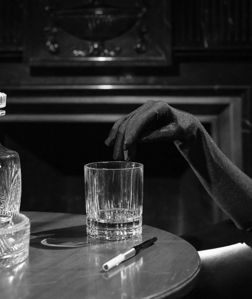 A gloved hand reaching towards a glass tumbler on a wooden table, with a crystal decanter and a pen, set against a dark, ornate fireplace background.